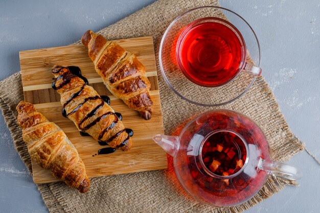 Croissants con salsa, té, tabla de madera sobre yeso y saco, plano.