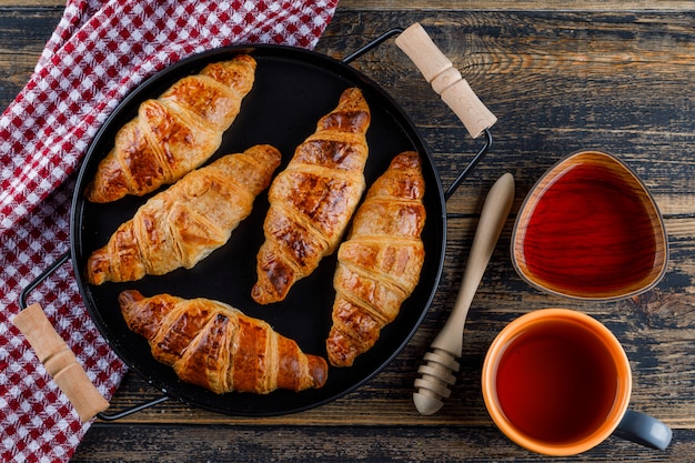 Croissants en una sartén con taza de té, miel, cazo plano sobre madera y papel de cocina