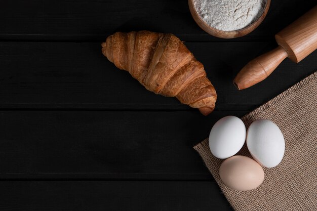 Croissants simples, cuenco de harina, rodillo y huevos crudos sobre superficie de madera oscura. Foto de alta calidad