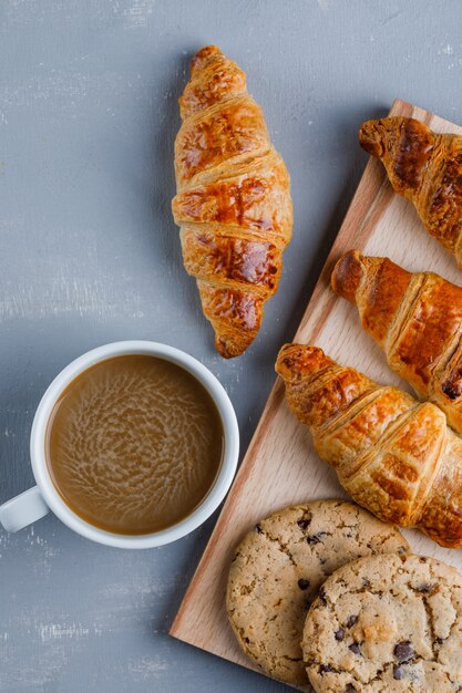 Croissants con taza de café, galletas planas sobre yeso y tabla de cortar