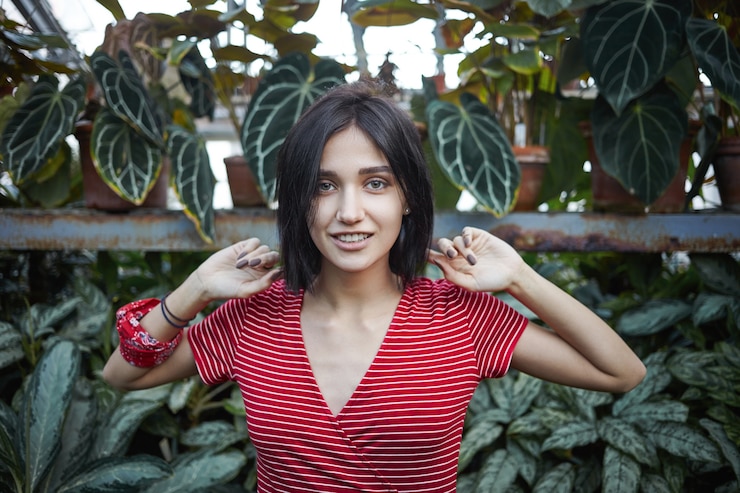 Photo Gratis cropped view of beautiful young european female with dark bib hairstyle relaxing outdoors, walking in jungle surrounded with exotic plants, feeling happy and carefree, smiling joyfully at camera
