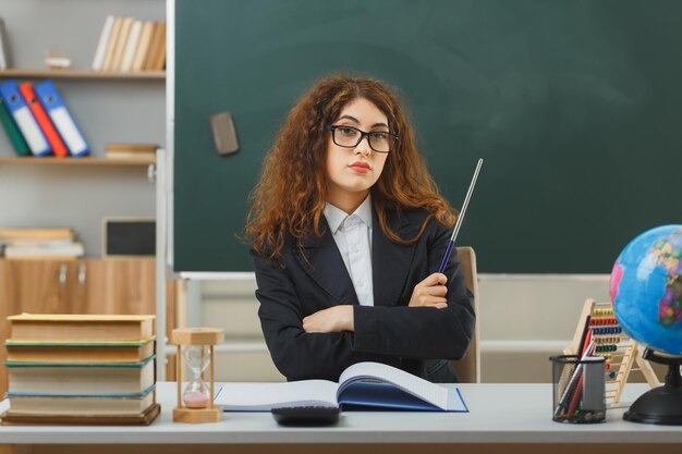 cruce estricto de manos joven maestra con gafas sosteniendo un puntero sentado en el escritorio con herramientas escolares en el aula