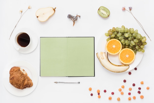 Cuaderno abierto en blanco con bolígrafo; cuerno; Café y frutas sobre fondo blanco