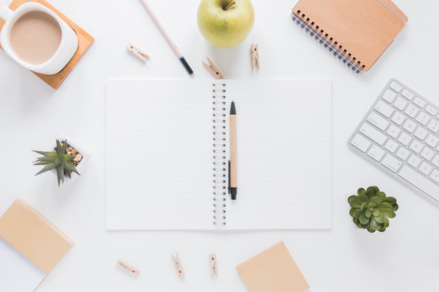 Cuaderno abierto con bolígrafo cerca de papelería y manzana en mesa blanca