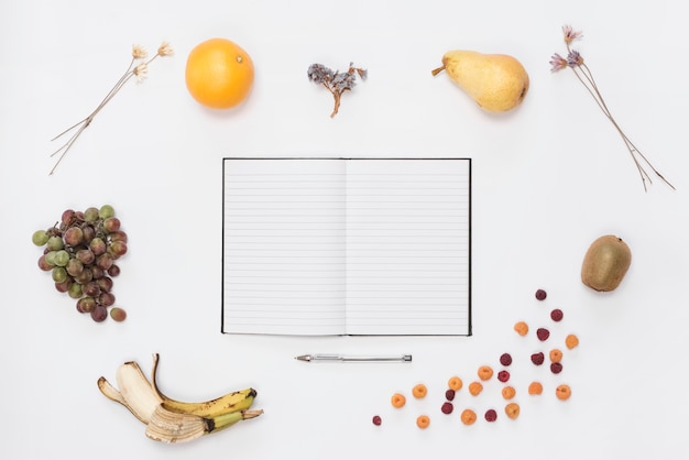 Un cuaderno abierto y un bolígrafo rodeado de frutas maduras sobre fondo blanco