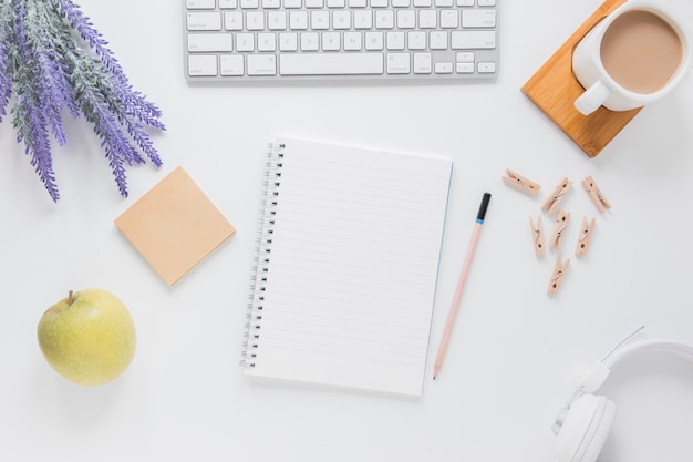 Cuaderno abierto cerca de papelería en mesa blanca con gadgets y taza de café