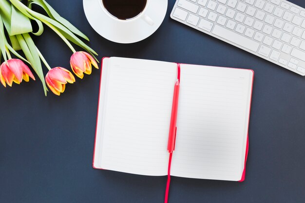 Cuaderno abierto cerca de la taza de café y tulipán en el escritorio con teclado