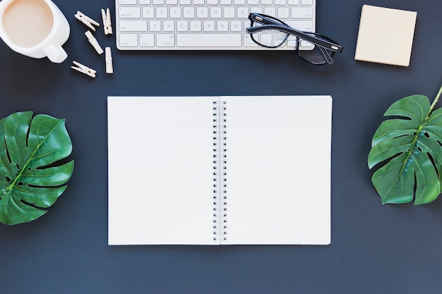 Cuaderno abierto cerca del teclado y la taza de café en el escritorio con hojas y vasos