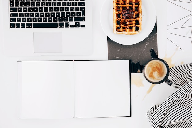 Cuaderno blanco en blanco; ordenador portátil; gofre; Taza de café y mantel sobre fondo blanco