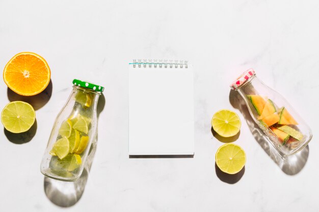 Cuaderno de bocetos vacío lima naranja y botellas de vidrio con cítricos cortados