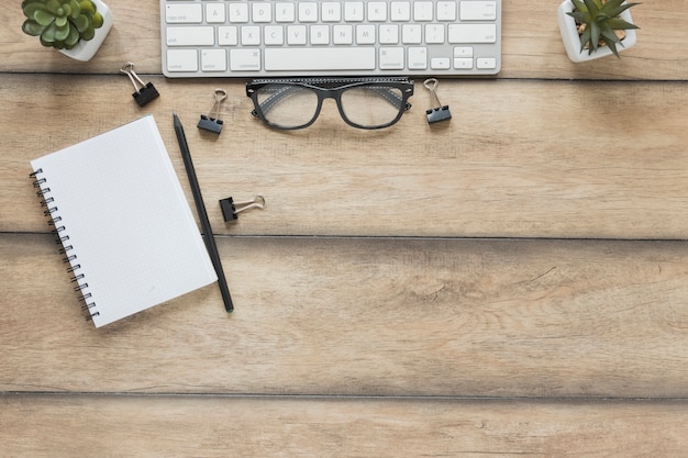 Cuaderno con bolígrafo colocado cerca del teclado y gafas en la mesa de madera