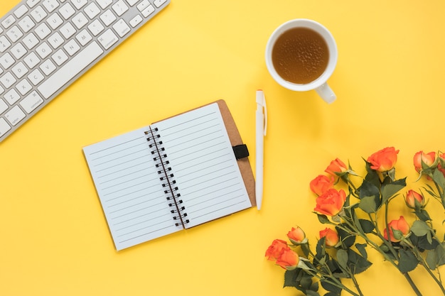 Cuaderno cerca de taza de bebida, teclado y rosas frescas con hojas verdes