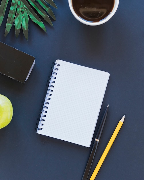 Cuaderno cerca de la taza de café y papelería en el escritorio con manzanas y palmeras