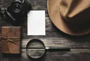 Foto gratuita cuaderno cerrado con tapa de cuero, hoja de papel blanco, sombrero marrón de fieltro, cámara y lupa grande aislada en mesa de madera envejecida negra