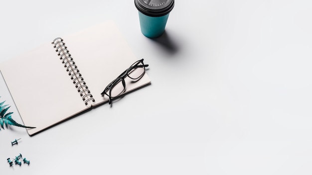 Un cuaderno de espiral abierto en blanco con gafas; Taza de café desechable y alfileres sobre fondo blanco