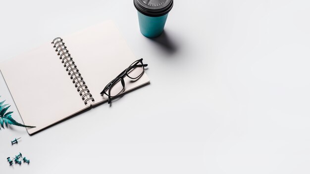 Un cuaderno de espiral abierto en blanco con gafas; Taza de café desechable y alfileres sobre fondo blanco