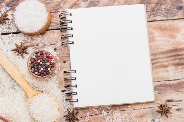 Foto gratuita cuaderno espiral en blanco; arroz crudo y especias secas sobre papel pintado de madera.