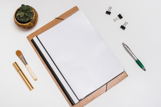 Cuaderno espiral en blanco con pincel de maquillaje; máscara; Clips de papel y pluma con planta en maceta en escritorio blanco