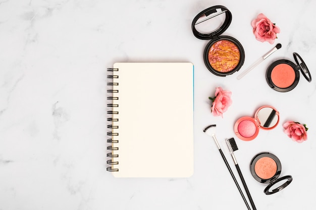 Cuaderno espiral en blanco con rosas rosadas; Pinceles de maquillaje y polvos compactos.
