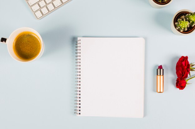 Cuaderno espiral en blanco con taza de té; teclado; planta de cactus; Rosas y lápiz labial sobre fondo azul