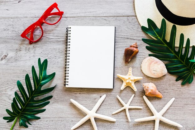 Cuaderno espiral con concha marina, hoja, sombrero de paja, gafas de sol y estrellas de mar en la mesa