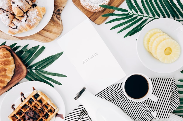 Cuaderno rodeado de croissant; waffles rodajas de piña; botella y taza de café sobre fondo blanco