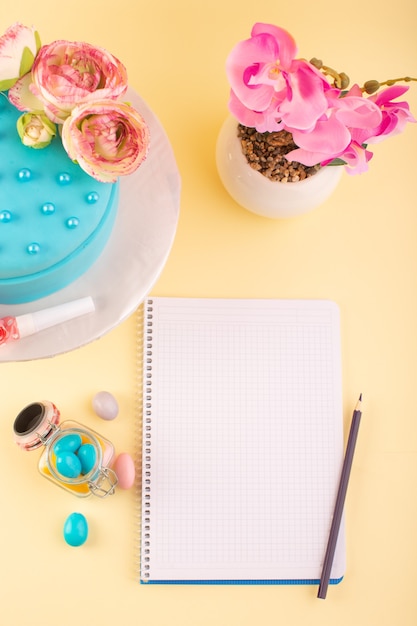 Un cuaderno de vista superior y un pastel con dulces y flores en la fiesta de celebración de cumpleaños del escritorio amarillo