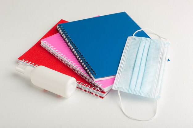 Cuadernos coloridos de vista frontal con spray y máscara sobre superficie blanca