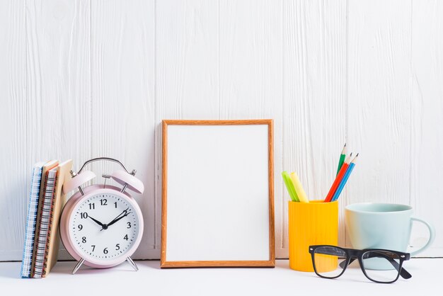 Cuadernos; despertador; marco en blanco porta lápices; Taza y anteojos contra papel pintado de madera blanco.