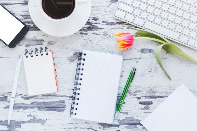 Cuadernos y taza de café cerca de teléfono inteligente y teclado en el escritorio con flor de tulipán