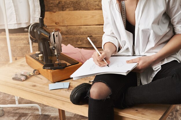 Cuando el hobby se convierte en trabajo real. Captura recortada de una creativa diseñadora de ropa femenina sentada en la mesa cerca de la máquina de coser en su taller, tomando notas o planeando un nuevo diseño para su línea de ropa