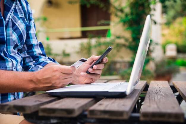 Cuarenta años de edad caucasian hombre mirando tarjeta de crédito mientras trabajaba en equipo portátil en terraza de jardín durante el día soleado de verano. Estilo de vida moderno - fin de semana del campo y concepto en línea de las compras.