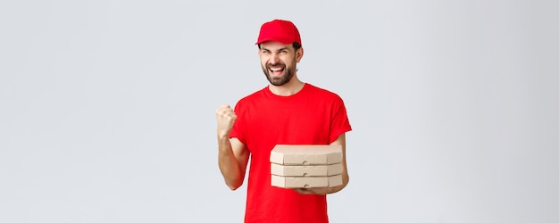 Cuarentena de entrega de alimentos quédese en casa y ordene el concepto en línea Mensajero entusiasta con camiseta roja y gorra que trae pedidos de pizza a la casa bomba de puño seguro de ganar celebrando la victoria