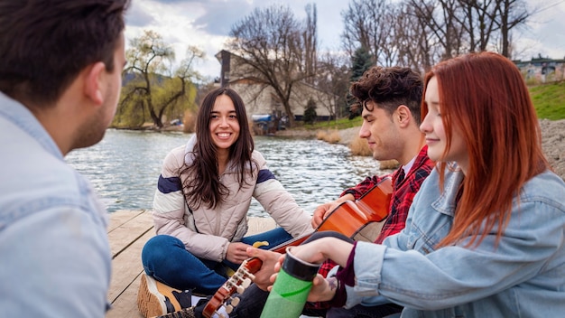 Cuatro jóvenes amigos cantando, descansando y tocando la guitarra cerca de un lago en un parque