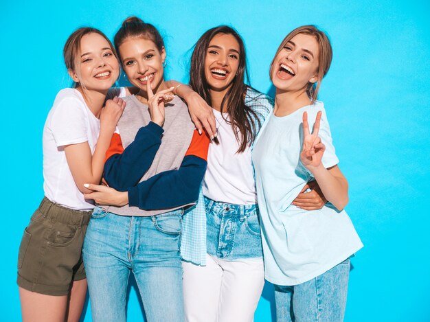 Cuatro jóvenes hermosas chicas hipster sonrientes en ropa de moda de verano. Mujeres despreocupadas atractivas que presentan cerca de la pared azul en estudio. Modelos positivos divirtiéndose y abrazándose