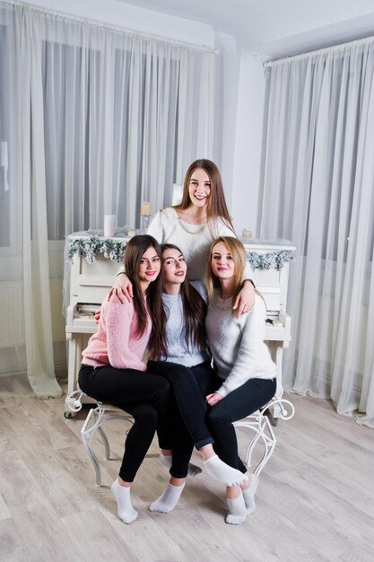 Cuatro lindas amigas usan suéteres cálidos y pantalones negros contra un viejo piano con decoración navideña en la habitación blanca