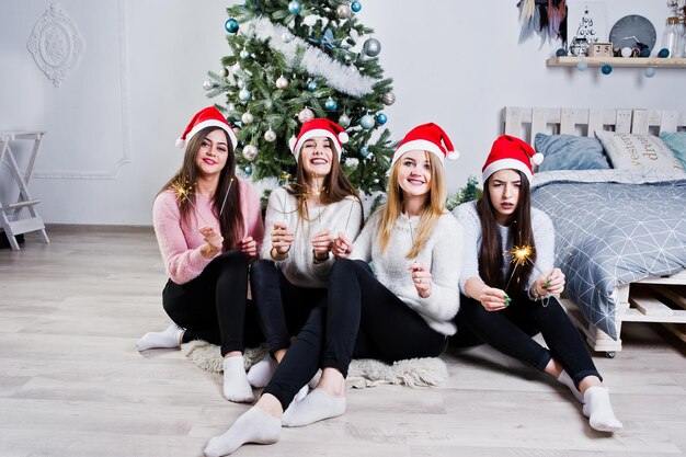 Cuatro lindas amigas usan suéteres cálidos, pantalones negros y sombreros rojos de santa contra el árbol de año nuevo con decoración navideña en la habitación blanca y sostienen luces de bengala en las manos