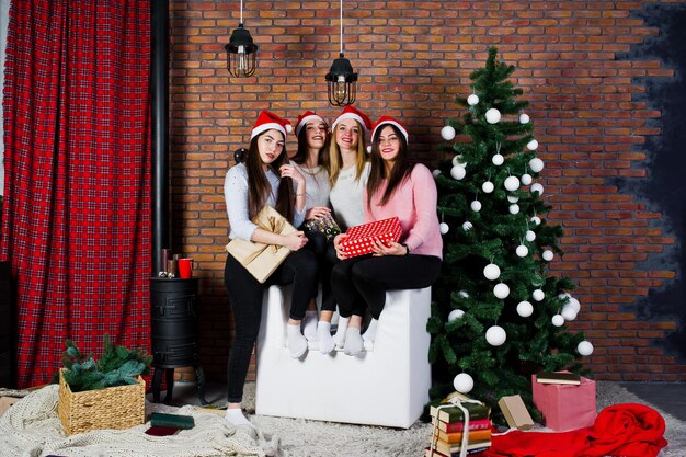 Cuatro lindas amigas usan suéteres cálidos, pantalones negros y sombreros de santa contra el árbol de año nuevo con decoración navideña en el estudio