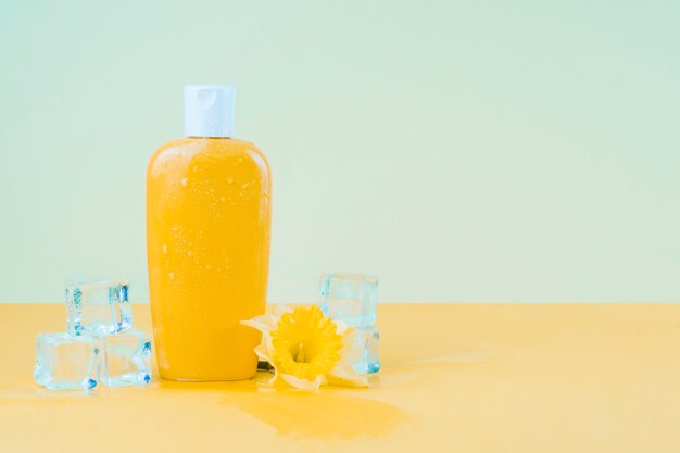 Cubitos de hielo de cristal con flor de narciso y botella de loción protectora amarilla contra el fondo verde