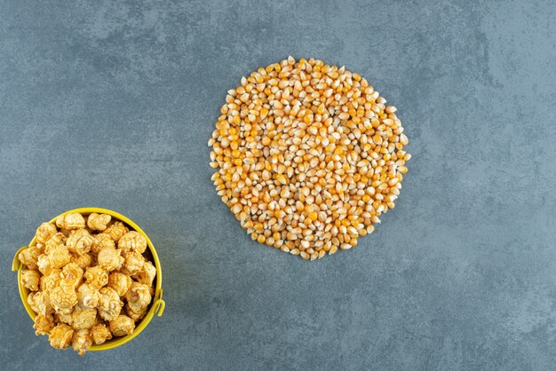 Cubo de dulces de palomitas de maíz junto a una pila redonda de grano de maíz sobre fondo de mármol. Foto de alta calidad