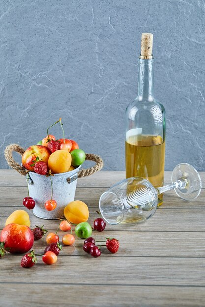 Cubo de frutas frescas de verano, botella de vino blanco y vaso vacío en la mesa de madera.