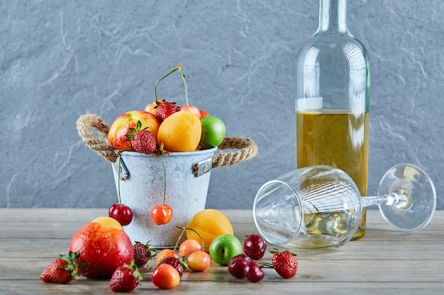 Cubo de frutas frescas de verano, botella de vino blanco y vaso vacío en la mesa de madera.