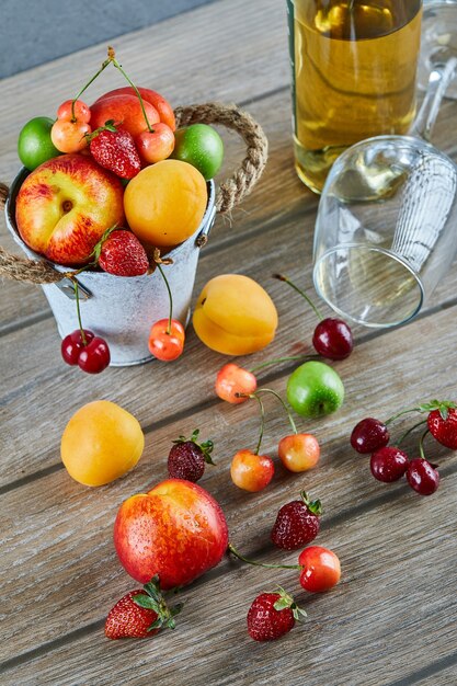 Cubo de frutas frescas de verano, botella de vino blanco y vaso vacío en la mesa de madera.