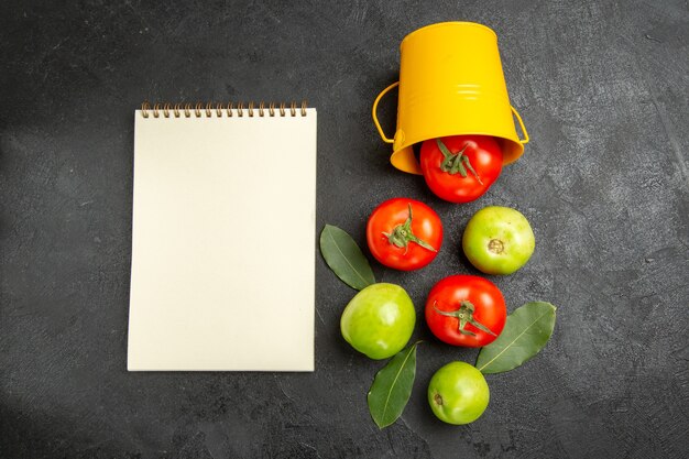 Cubo de vista superior con hojas de laurel de tomates rojos y verdes y un cuaderno sobre fondo oscuro