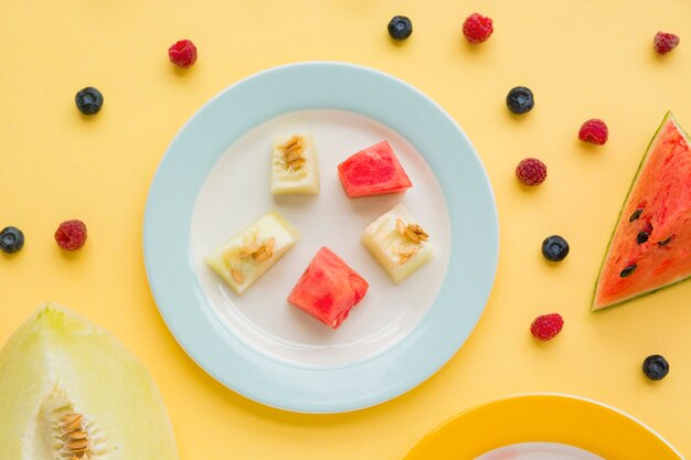 Cubos de melón y sandía en un plato con frambuesas y arándanos sobre fondo amarillo