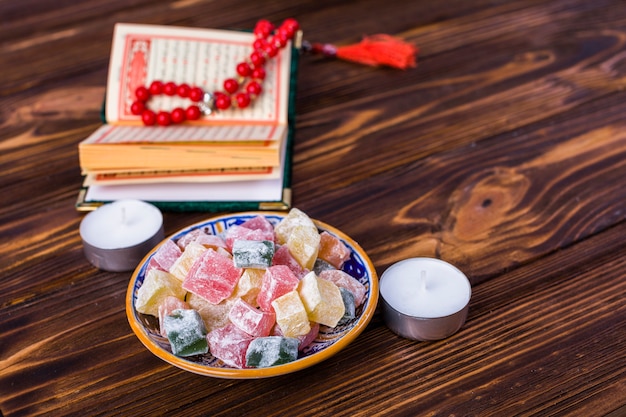 Foto gratuita cubos multicolores de rakhat-lukum en placa con libro sagrado islámico; rosario y velas en el escritorio de madera