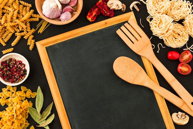 Cuchara de madera y espátula sobre pizarra rodeada de ingredientes de pasta.