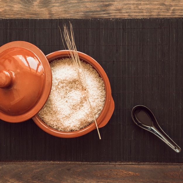 Cuchara negra con una olla de granos de arroz crudo con tapa en la estera de lugar sobre la mesa de madera