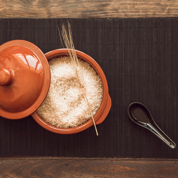 Cuchara negra con una olla de granos de arroz crudo con tapa en la estera de lugar sobre la mesa de madera