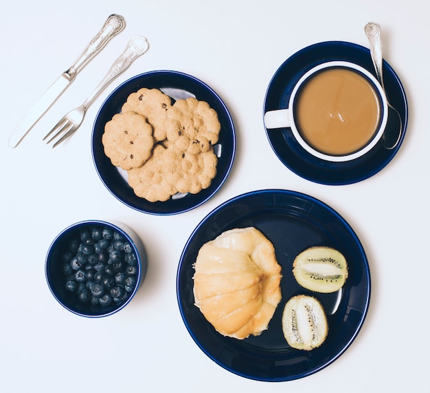 Cuchillería; galletas; kiwi; arándanos; Taza de café y pan sobre fondo blanco